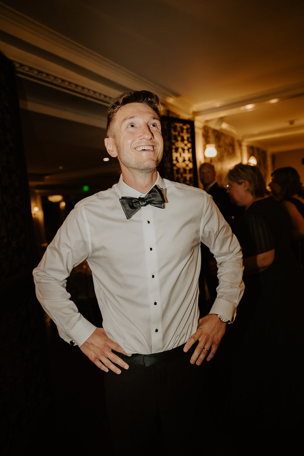 Groom stands with hands on his hips, looking up with a joyful smile during the reception.